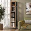 A tall, narrow bookshelf filled with books and decorative items, placed next to a window with sheer curtains and a green sofa with a yellow throw blanket. Anteprima dell’immagine prodotto
