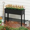 A raised garden bed filled with flowers, placed on a wooden deck next to a white wall. Gardening tools and a watering can are also visible. Voir les images du produit