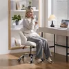 A woman is sitting in a white office chair, drinking from a cup, in a home office setting with a bookshelf and desk. Produktbild-Vorschau