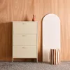 A cream-colored metal shoe cabinet with three drawers is placed against a wooden wall. To the right, there is a decorative arched panel and a striped stool. A small red vase sits on top of the cabinet. Produktbild-Vorschau