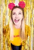 A woman wearing mouse ears and a yellow shirt with text, sticking her tongue out, in front of a gold fringe curtain. Produktbild-Vorschau