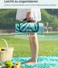 A person holding a folded picnic blanket in a grassy outdoor setting. Produktbild-Vorschau