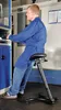 A man in a blue lab coat is sitting on an adjustable stool in what appears to be a workshop or laboratory. Produktbild-Vorschau