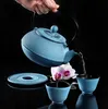 A hand pours water from a blue cast iron teapot into a blue cast iron teacup, with a pink orchid in the background. Náhled obrázku produktu