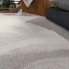 A close-up view of a textured, light gray carpet with subtle wave-like patterns. In the background, a woven basket and dark gray cushions are visible. Produktbild-Vorschau