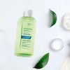 A bottle of shampoo is laid out on a white surface with some green leaves and a loofah. Produktbild-Vorschau