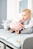 A baby is sitting on a changing mat with two plush toys. Anteprima dell’immagine prodotto