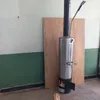 A stainless steel water heater with a chimney pipe and a shower head attachment stands in front of a wooden board against a wall. Produktbild-Vorschau