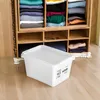 A white storage box with a lid is placed on a wooden floor in front of a wooden shelf unit filled with folded clothes. Produktbild-Vorschau