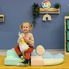 A young girl sits on a set of colorful soft play blocks, holding a stuffed bunny. Produktbild-Vorschau