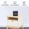 A black cube clock with white digital display sits on a light wood and white nightstand. Produktbild-Vorschau