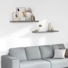 Two gray floating shelves are mounted on a white wall above a light gray sectional sofa. The shelves are decorated with various decorative items, including vases, books, and framed pictures. Produktbild-Vorschau