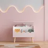 A pink toy box with a lid open, displaying books and toys, in a child's room with a wavy light fixture on the wall. Náhled obrázku produktu