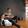 A man is playing an electric guitar while sitting on a bean bag chair in a room with a brick wall. Voir les images du produit