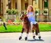 A young girl with red hair wearing a flower crown rides a brown toy horse with white leg wraps in a park setting. Anteprima dell’immagine prodotto