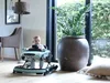 A baby is sitting in a car-shaped baby walker in a room with large windows and plants. Produktbild-Vorschau