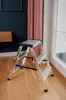 A silver step stool with three steps is placed on a wooden floor in a room with dark blue walls and pink curtains. Produktbild-Vorschau