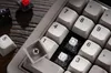 A close-up view of a mechanical keyboard with several keycaps removed, revealing the switches underneath. Some loose keycaps are scattered around the keyboard. Produktbild-Vorschau