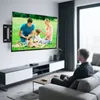A man is sitting on a couch watching a TV mounted on the wall. The TV is displaying a family picnic scene. A media console is below the TV. Produktbild-Vorschau