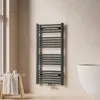 A modern bathroom setting with a dark grey towel radiator mounted on a beige wall, next to a white bathtub and a wooden stool with a folded towel. Anteprima dell’immagine prodotto