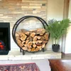 A round metal firewood rack filled with logs sits next to a fireplace, with a potted plant to the right. Náhľad obrázku produktu