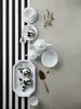A collection of white porcelain tableware and desserts arranged on a gray surface with a black and white striped background. Produktbild-Vorschau