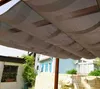 A view from underneath a brown mesh pergola canopy, looking out at houses and a stone wall. Podgląd zdjęcia produktu