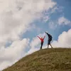 Two people are standing on a grassy hill with their arms raised in celebration against a cloudy blue sky. Produktbild-Vorschau