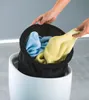 Hands are shown placing a black laundry bag filled with blue and yellow towels into a white cylindrical container, possibly a stool or hamper. Podgląd zdjęcia produktu