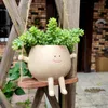 A cute, smiling planter pot shaped like a round character with arms and legs is hanging from a rope swing. It is filled with green succulents and is set against a blurred background of a house and greenery. Anteprima dell’immagine prodotto