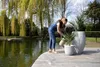 A woman watering plants on a wooden deck next to a pond with weeping willow trees in the background. Voir les images du produit