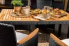 A dining set with a wooden table and rattan chairs, set up on a patio with food and drinks. Náhled obrázku produktu