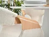 Close-up of a chair armrest with a textured fabric and a wooden curved support, with a table and greenery in the background. Produktbild-Vorschau