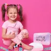 A young girl with pigtails is sitting on a pink background, showing off her painted nails. Around her are items from a makeup and nail cosmetic set for children. Produktbild-Vorschau