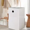 A white dehumidifier is placed on a rug in a room with clothes hanging in the background and a wooden stool to the side. Produktbild-Vorschau
