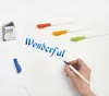 A hand is writing the word "Wonderful" with a blue calligraphy pen on white paper. Several other calligraphy pens with colored caps are arranged on the paper. Voir les images du produit