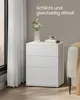 A white bedside table with three drawers, decorated with a vase of flowers and books, placed next to a window with sheer curtains. Produktbild-Vorschau