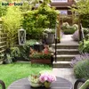 A lush, green garden with a stone pathway leading up to a house, featuring various plants, flowers, and decorative elements. Anteprima dell’immagine prodotto