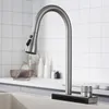 A kitchen faucet is shown in a close-up view, positioned over a sink. The background features a tiled wall and a countertop with some blurred items. Podgląd zdjęcia produktu