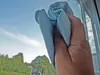 A hand holding a blue cleaning cloth, cleaning a window with a view of trees and sky. Voir les images du produit