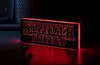 A Stranger Things logo light is displayed on a dark surface, with a hint of a retro television or computer monitor in the background. Produktbild-Vorschau
