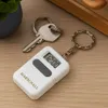 A white keychain with a digital clock and the name MARSCHALL is shown on a wooden surface next to a key and a plant. Produktbild-Vorschau