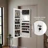 A white jewelry cabinet with a mirror is mounted on a door. The cabinet is open, displaying various compartments filled with jewelry. A small inset shows a close-up of the lock and keys. Náhľad obrázku produktu