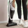 A person is holding a white and black vacuum cleaner while standing on a wooden floor. Voir les images du produit