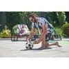 A man is kneeling on a wooden deck next to a swimming pool, holding a yellow and black device near a soccer ball. Another person is relaxing on a lounge chair in the background. Produktbild-Vorschau