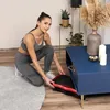 A woman in workout clothes is placing a vibration plate into a storage compartment under a blue coffee table. Náhled obrázku produktu