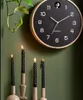 A black Karlsson wall clock with a wooden frame hangs on a green wall above four lit black candles in beige holders. Produktbild-Vorschau
