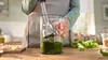 A person is using an immersion blender to make pesto in a glass beaker on a wooden countertop. Produktbild-Vorschau