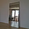 A tall, rectangular mirror with a geometric design, leaning against a wall in a room with wooden flooring. The mirror reflects a window, a chair, and a vase with dried plants. Náhled obrázku produktu