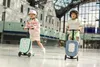 Two children are riding scooters with attached suitcases in a large indoor space, possibly an airport or train station. Produktbild-Vorschau